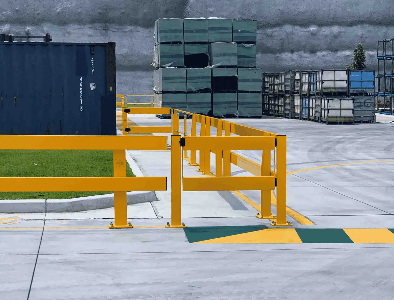 Yellow industrial safety barriers and a swinging gate installed in an outdoor shipping area with storage containers and stacked pallets in the background.