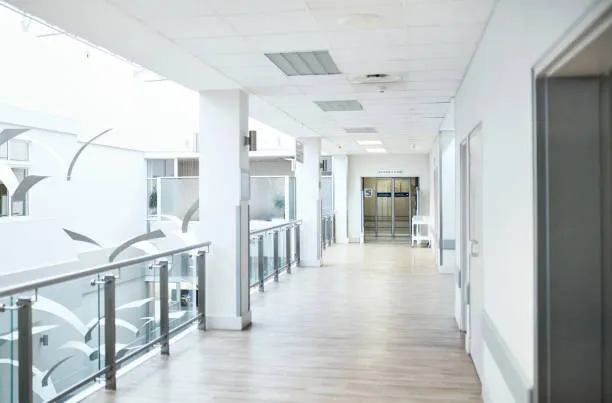 A bright, modern hospital or clinic corridor featuring light wood-look flooring, white walls, and a glass safety railing