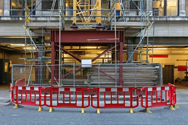A construction site area enclosed by red and white pedestrian safety barriers and tall wire mesh fencing