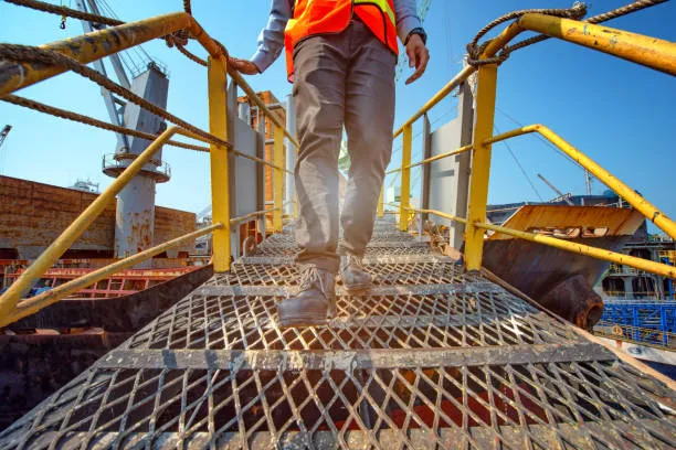 A low-angle view of a worker in a safety vest walking down a metal grate gangway with yellow handrails