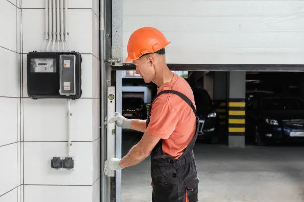 A technician in an orange hard hat and work overalls using a spirit level to check the alignment of an automatic garage door track