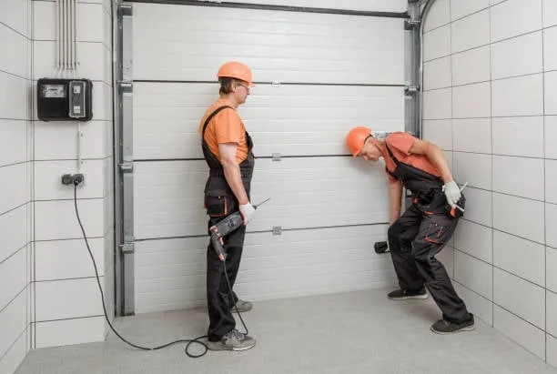 Two technicians in orange hard hats and dark work overalls installing or repairing a white sectional garage door in a tiled service area.
