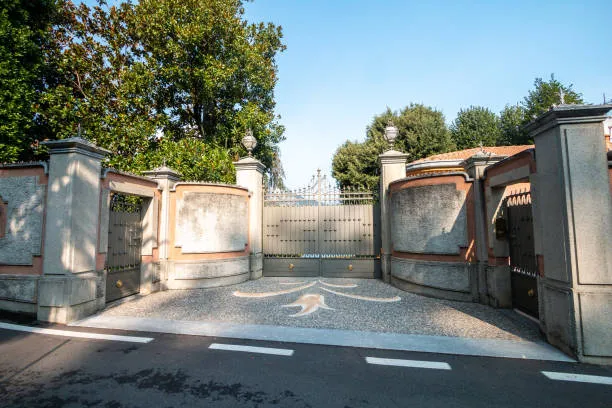 An elegant, grey metal driveway gate and matching pedestrian side gates set into a curved masonry wall with stone pillars and decorative finials