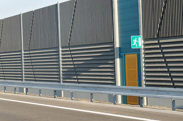 A section of a tall, grey sound-insulating wall along a highway featuring a bright orange emergency exit door