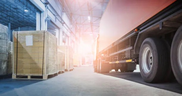 A large orange semi-truck parked next to a row of shrink-wrapped pallets at a warehouse loading dock