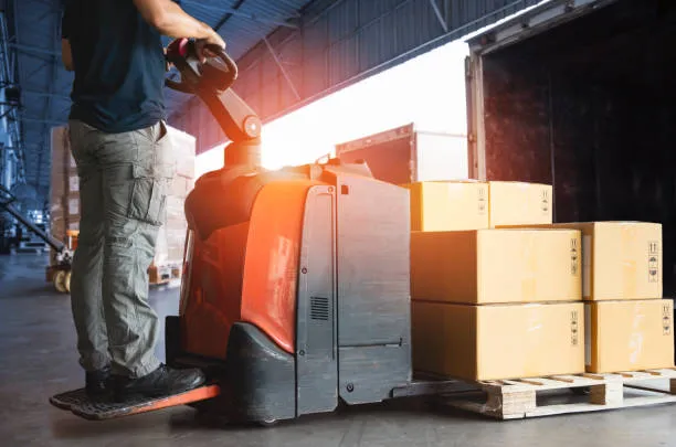 A worker operating an electric pallet jack loaded with cardboard boxes