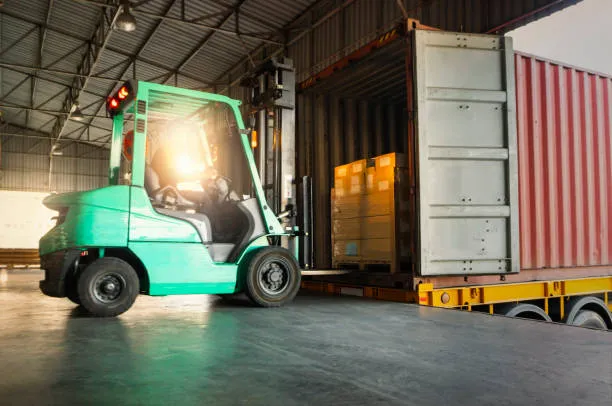 A green forklift operator loading a pallet of goods into a large red shipping container at a warehouse loading dock during sunset.