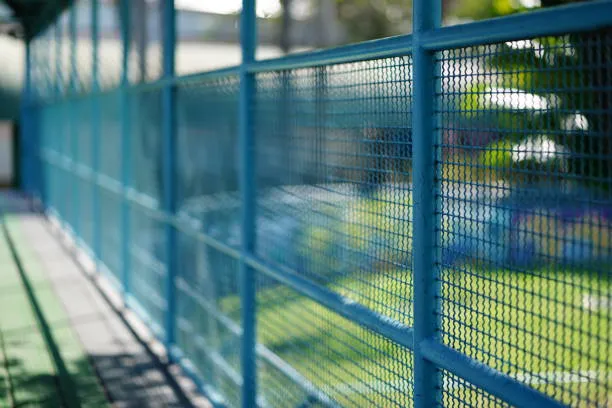 A close-up, angled view of a blue metal mesh fence providing a secure boundary for an outdoor sports court or park area.