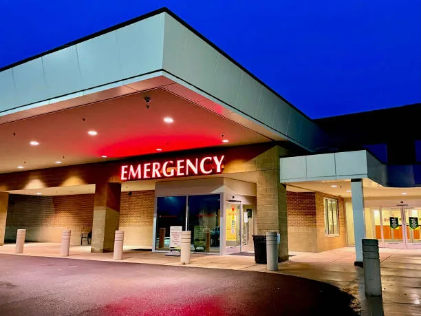 An exterior view of a hospital emergency room entrance at dusk, featuring a large, illuminated red 'EMERGENCY' sign