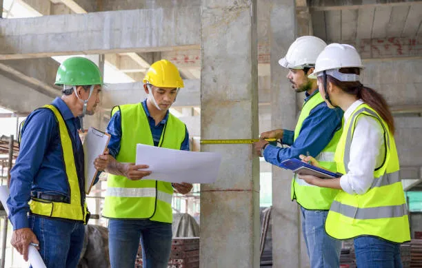 A team of four construction professionals in hard hats and high-visibility vests reviewing blueprints and using a yellow measuring tape to inspect a concrete pillar at a building site.