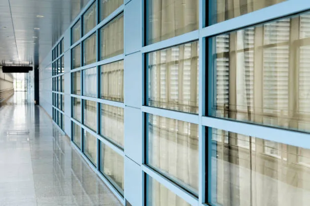 A perspective view of a long, bright office corridor featuring a polished floor and a wall made of light blue framed glass partitions
