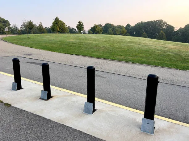 Four black, heavy-duty bollards installed in a concrete base along the edge of a paved road