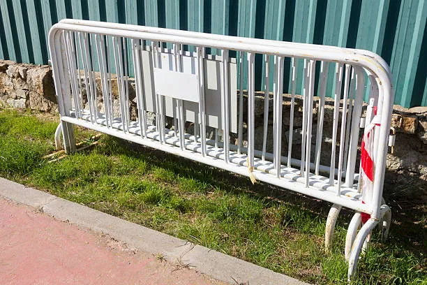 A row of white metal crowd control barriers stacked together on a patch of grass next to a paved walkway