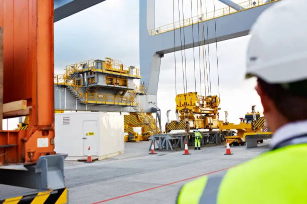 A port worker in a high-visibility vest and white hard hat overseeing the operation of a massive ship-to-shore crane